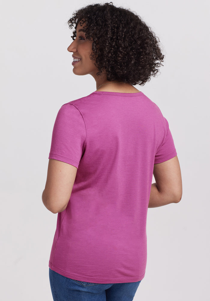 A person with curly hair is smiling and facing away from the camera, wearing the Woolx Liza Crew Neck Tee in Purple Wine and blue jeans, standing against a plain light background.