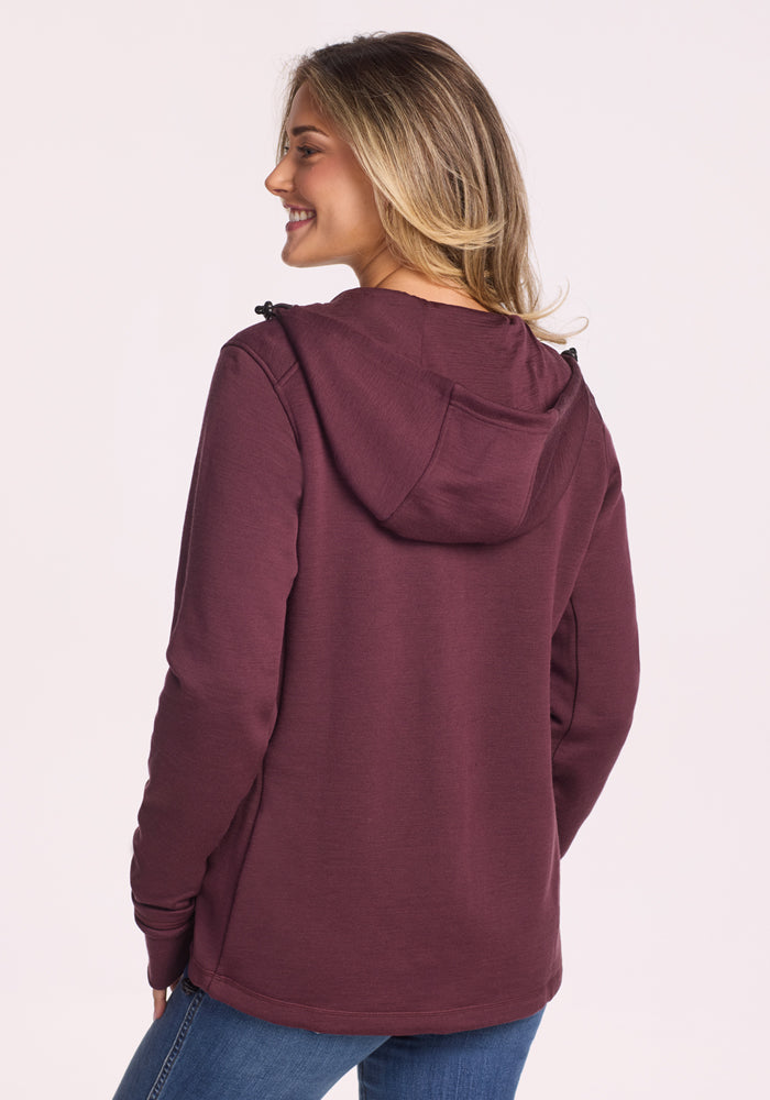 A person with long, light brown hair is smiling and wearing the Woolx Cubby Hooded Sweatshirt in Grape Harvest with blue jeans, shown from the back against a light background.