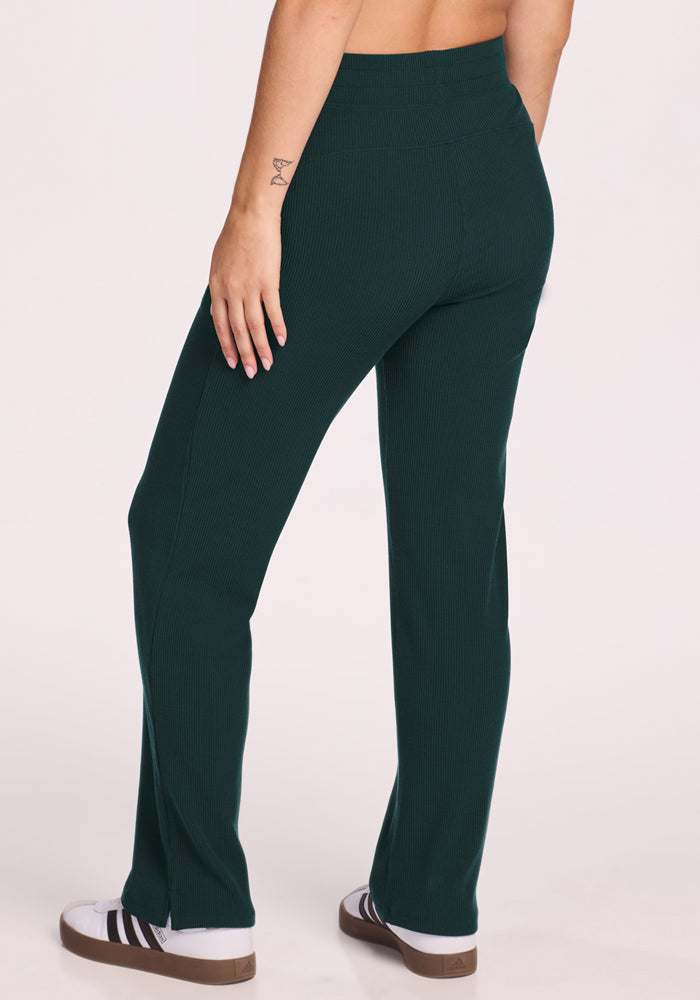 Wearing Woolx's Luca Ribbed Pants in Deep Timber, a person stands with their back to the camera, paired with white sneakers featuring black stripes. A small tattoo appears on their left forearm.