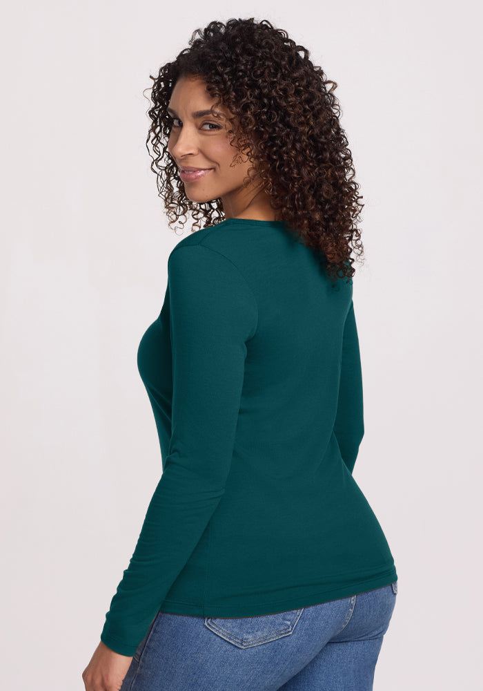 A woman with curly hair wears the Woolx Layla V Neck Top in Riverbank Green and blue jeans, standing against a plain light background, looking back over her shoulder and smiling.