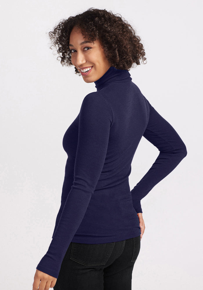 A woman with curly hair smiles over her shoulder, wearing the Woolx Sage Ribbed Turtleneck in Midnight Sapphire and black jeans, set against a light background.