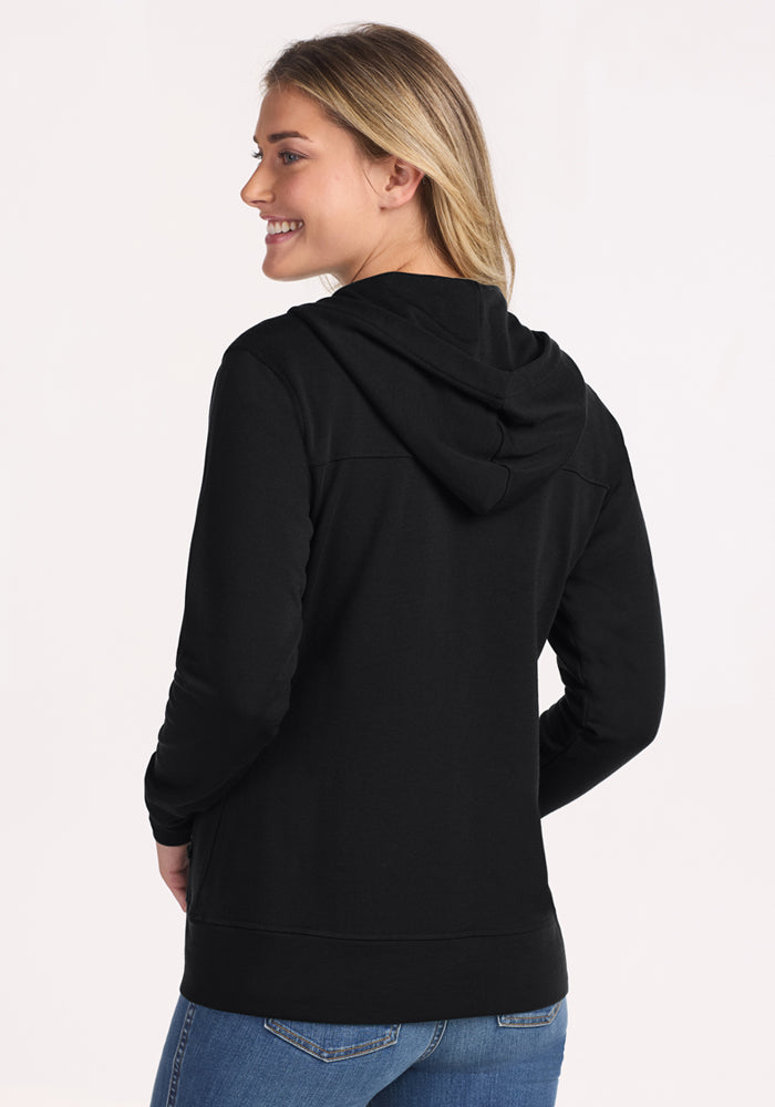 A woman with long blonde hair is smiling and facing away from the camera, wearing the Woolx Darby Zip Up Hoodie in black and blue jeans against a light background.