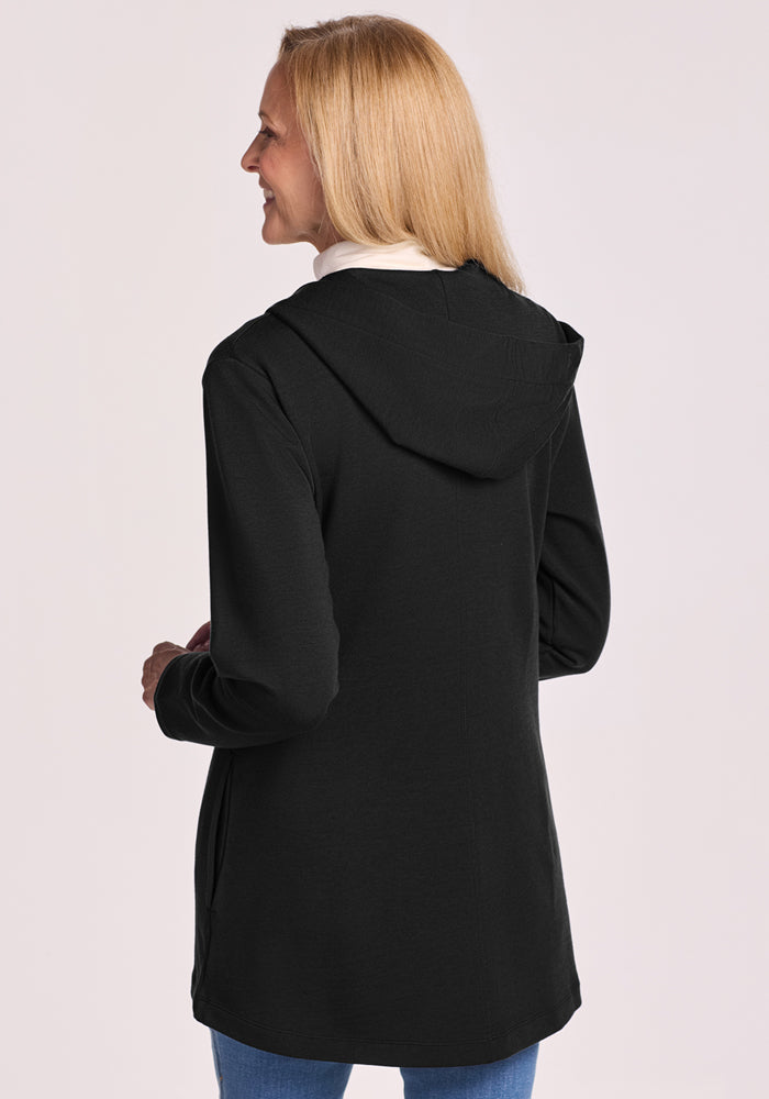 A woman with straight blonde hair, seen from the back, wears the Woolx Edna Hooded Cardigan in black over a white top and blue jeans, standing against a plain light background.