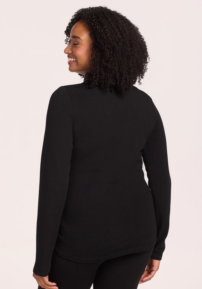 A woman with curly hair smiles, facing away from the camera in a Woolx Sadie Baselayer Top - Black and black pants, set against a light background.
