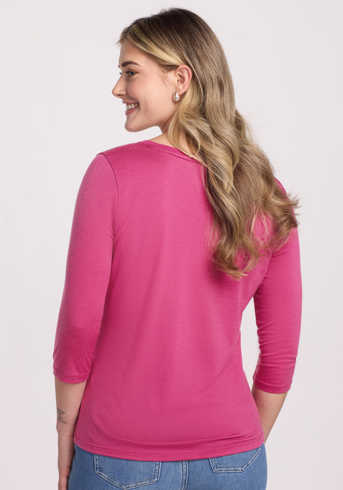 A woman with long, wavy blonde hair smiles as she faces away from the camera, wearing blue jeans and the Woolx Jenny 3/4 Sleeve Crew Neck in Raspberry Fizz. The background is plain and light-colored.