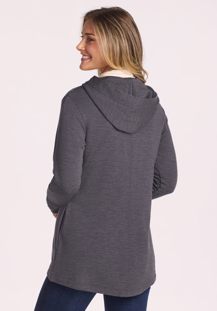 A woman with blonde hair smiles while wearing the Woolx Edna Hooded Cardigan in Cinder Melange over a white top and blue jeans. She is turned slightly to the side, facing away from the camera against a plain light background.