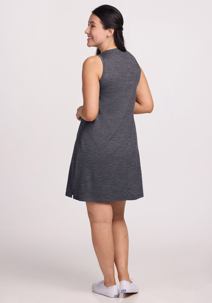 A woman with long dark hair, wearing the Woolx Clara Dress in Cinder Melange and white slip-on shoes, stands turned slightly to the side and smiles against a plain light background.