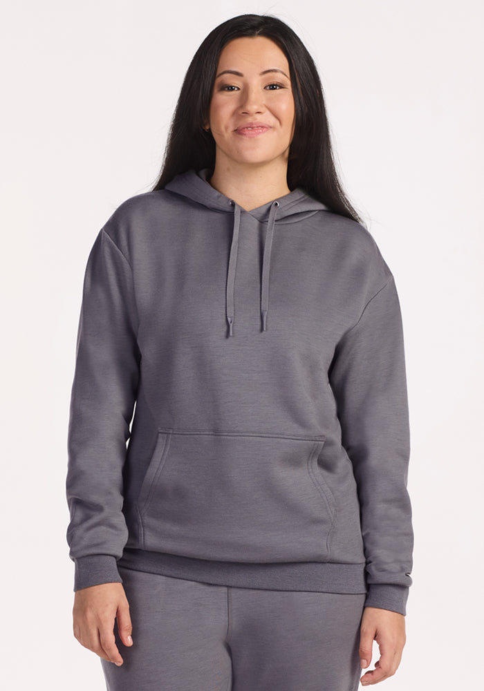 A woman with long dark hair smiles while wearing the Woolx Avery Hoodie in Arctic Dusk, made from recycled polyester. She stands against a plain light background with her hands relaxed at her sides. 