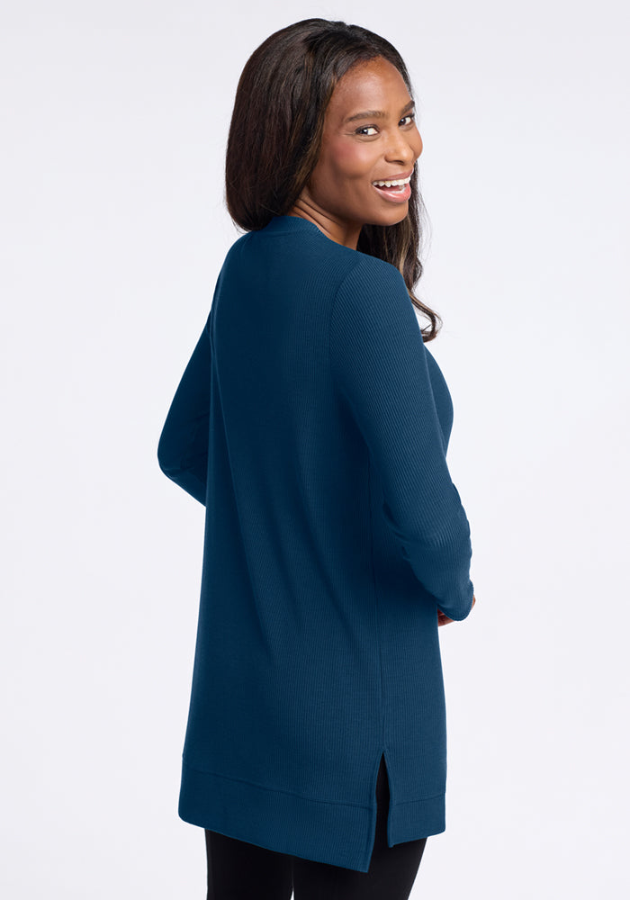 A woman with long dark hair smiles over her shoulder, wearing the Woolx Evie Ribbed Tunic in Majolica Blue and black pants. The temperature-regulating Merino wool keeps her comfortable all day against a plain white background.