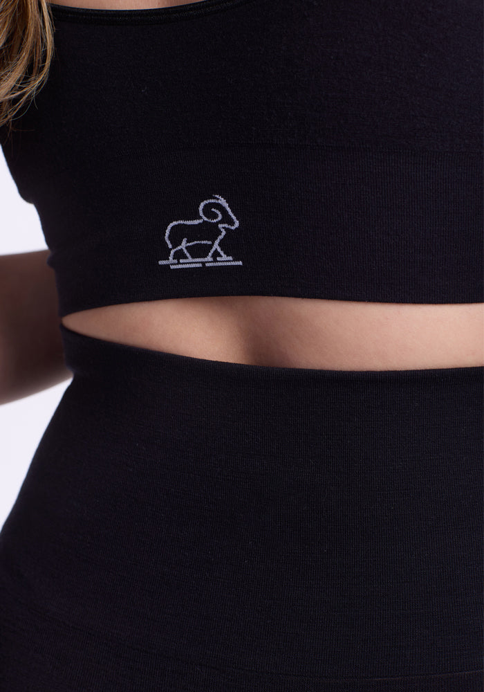 Close-up of a person wearing Woolx Ellis Seamless Petite Tights in black, featuring tummy control and paired with a matching black, sleeveless merino crop top that sports a small white ram logo and reveals the midriff.
