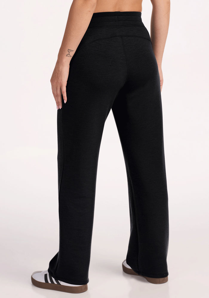 A person is shown from the waist down wearing Woolx Bree Lounge Pants in black and black-and-white sneakers. Their left arm has a small visible tattoo, and the background is plain and light-colored.