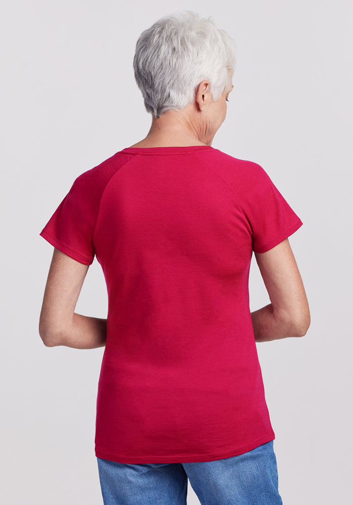 An older woman with short gray hair, seen from behind, wears the Woolx Mia V Neck shirt in Poppy Spark and blue jeans against a plain light gray background.