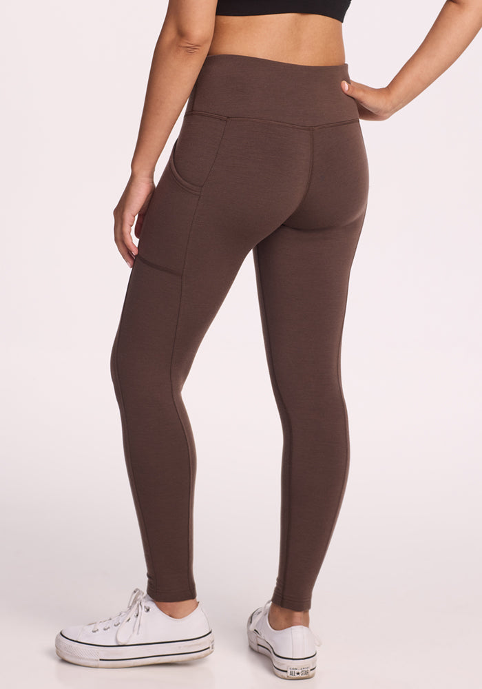 A person stands with one hand on their hip, shown from the back, wearing Woolx Piper Pocket Leggings in Bracken and white sneakers. The high-waisted, moisture-wicking leggings feature side pockets. The background is plain and light-colored.