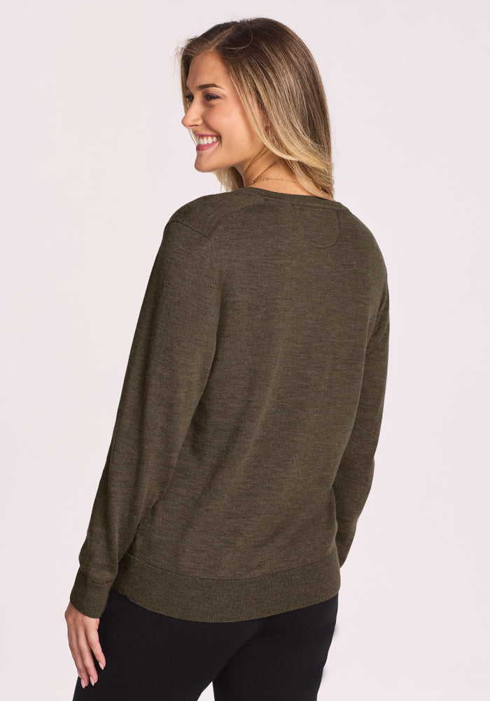 A woman with long, light brown hair smiles, standing slightly turned toward the camera. She wears the Woolx Francesca Sweater in Pine Needle and black pants against a plain light background.