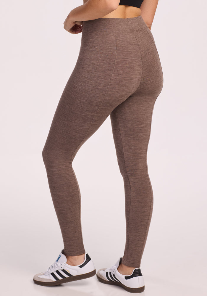 A person models Woolx Stella Leggings in Simply Taupe, a high-waisted brown style, paired with black-and-white sneakers, standing mid-torso down against a plain light background.