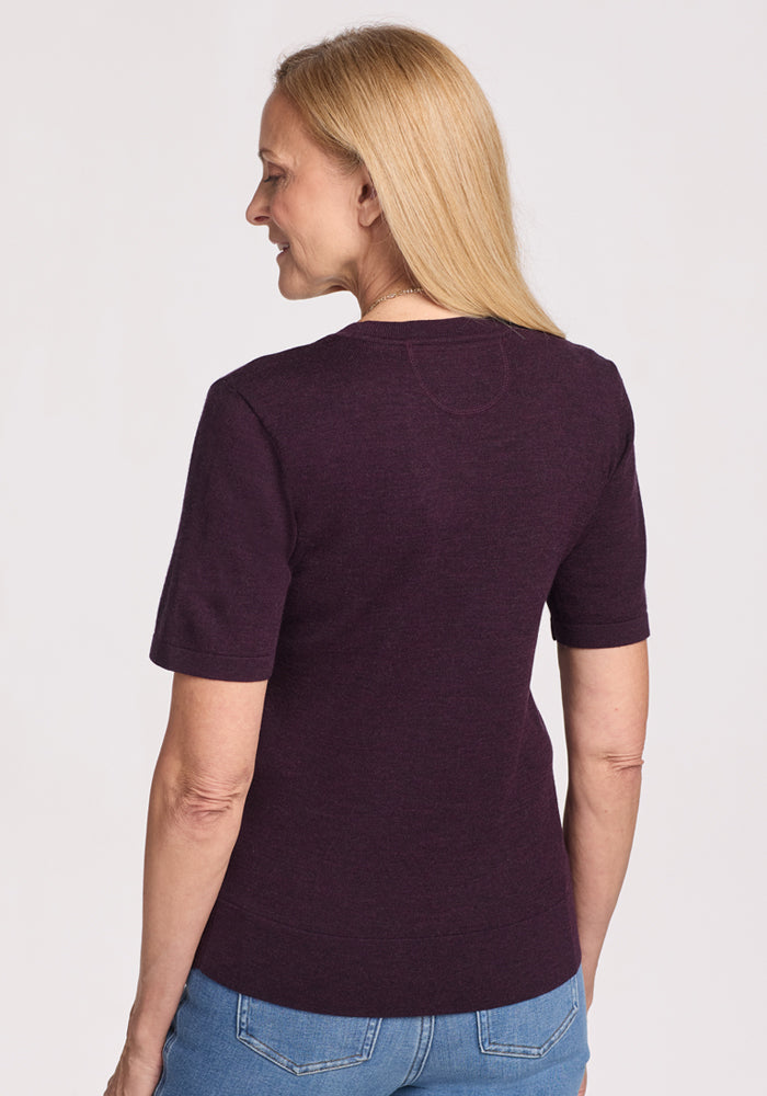 A woman with long blonde hair faces away, wearing the Woolx Anna Crew Sweater in Deep Plum and blue jeans against a plain light background.