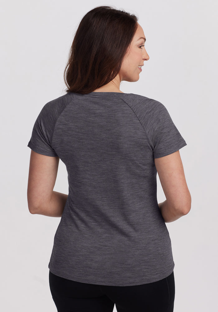 A woman with long brown hair is seen from behind, wearing the Woolx Mia V Neck - Cinder Melange short-sleeved tee and black pants against a plain light background.