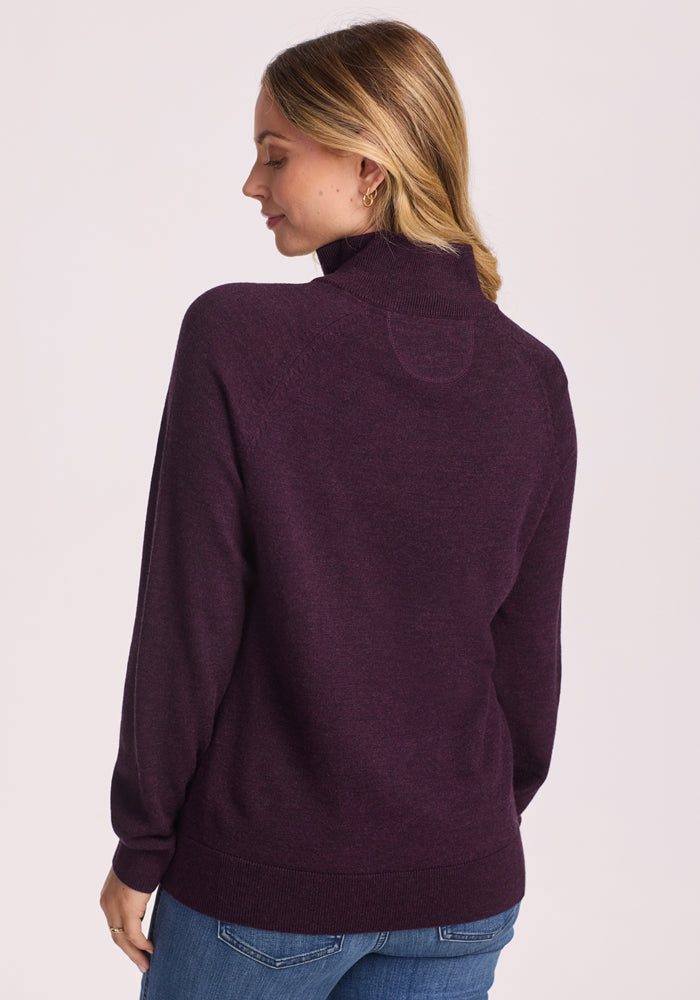 A woman with blonde hair, wearing the Woolx Collins Half Zip Sweater in Deep Plum and blue jeans, stands facing away and looks over her shoulder with a slight smile against a plain light background.