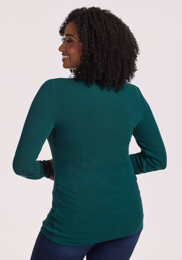 A woman with curly hair wears the Woolx Reese Ribbed Henley in Riverbank Green and blue jeans, smiling over her shoulder with her back to the camera against a light background.