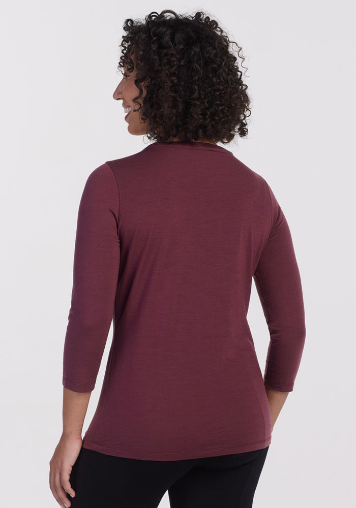 A woman with curly hair models the Woolx Jenny 3/4 Sleeve Crew Neck in Grape Harvest, standing with her back to the camera and wearing black pants against a plain light background.
