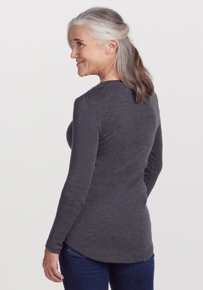 Older woman with gray hair pulled back, wearing the Woolx Eva Tunic in Cinder Melange and blue jeans, smiles over her shoulder against a plain white background—a cozy winter essential.