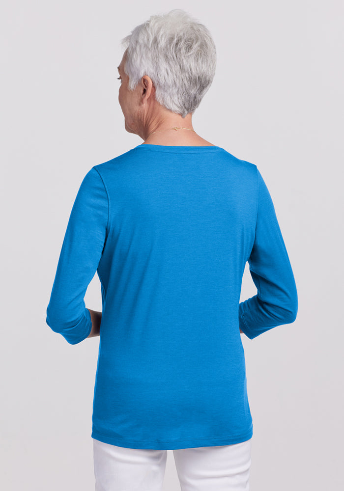 An older woman with short gray hair is shown from the back, wearing a bright blue Woolx Elena 3/4 Sleeve V Neck in Wave Rider and white pants against a plain light background.