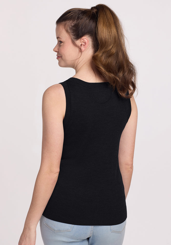 A woman with long brown hair in a ponytail faces away, wearing the Woolx Chessy Tank Top in black and light blue jeans—a perfect layering piece—against a plain light background.
