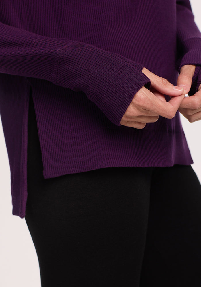 A person is shown from the waist down, wearing the Woolx Ainsley Sweater in Majestic Purple—featuring ribbed Merino wool and a side slit—paired with black leggings, with hands gently touching.