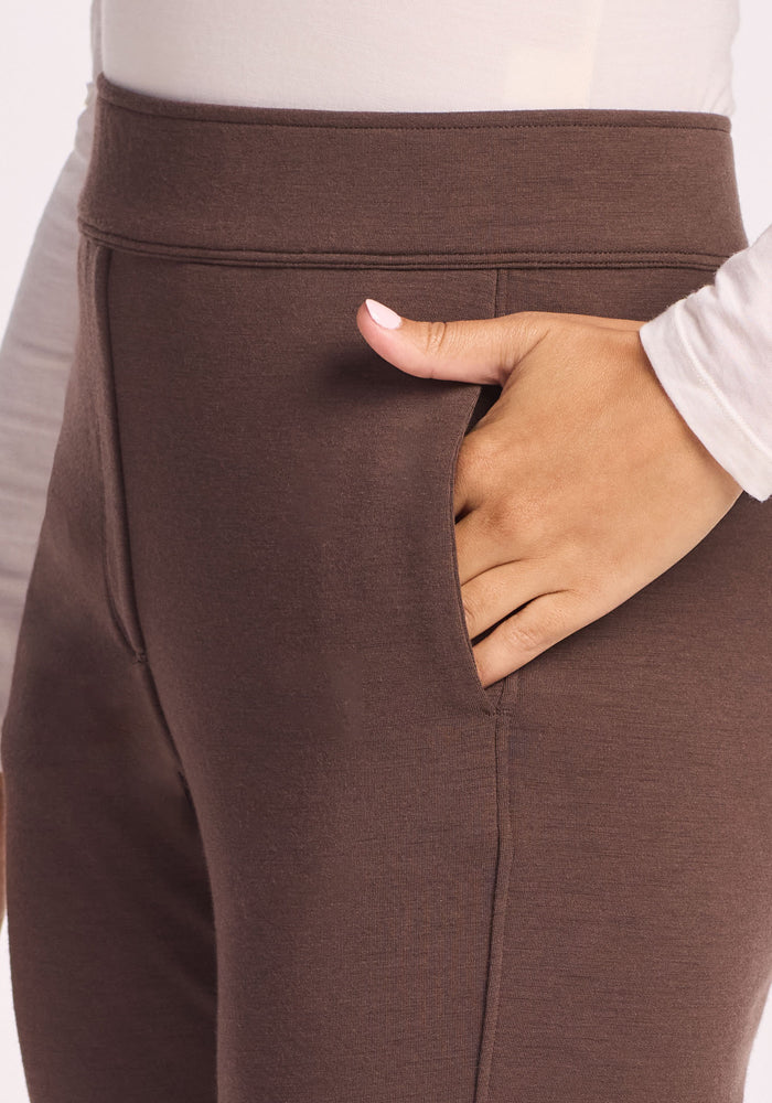 Close-up of a person wearing Woolx Emerson Straight Leg Pants in Bracken and a white long-sleeve shirt, with their right hand in the pants pocket. The background is plain and light-colored.