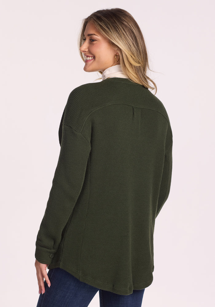 A woman with long, blonde hair smiles over her shoulder, wearing the Woolx Oaklynn Waffle Shacket in Driftwood Fern and dark blue jeans. The shacket’s cozy texture pops against a plain, light-colored background.