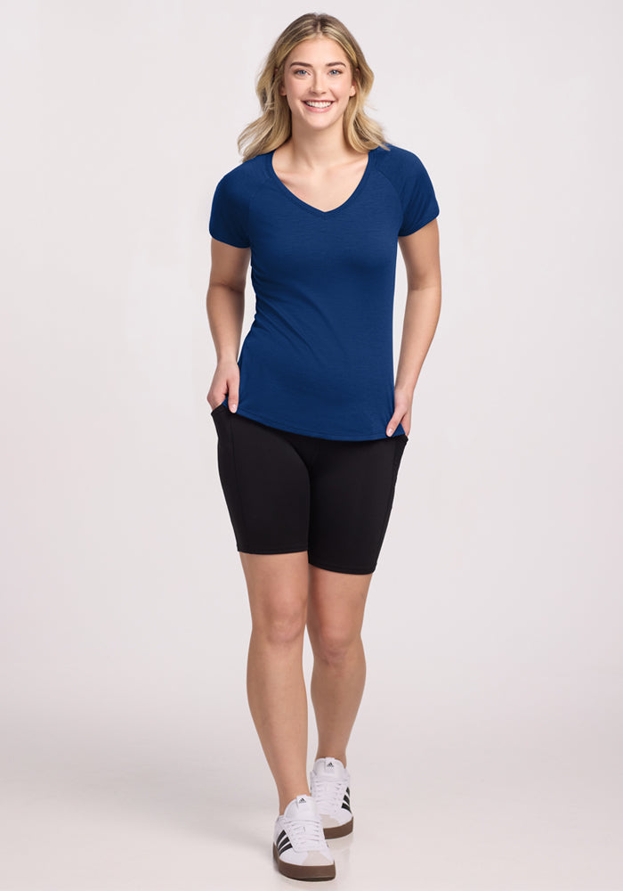 A blonde woman smiles with her hands in her black shorts' pockets, wearing the Woolx Mia V Neck in Estate Blue and white sneakers. The background is plain and light-colored.