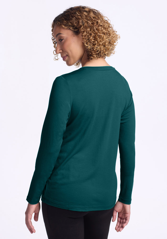A person with short curly hair is shown from the back, wearing the Woolx Millie Long Sleeve in Riverbank Green and black pants, standing against a plain white background.