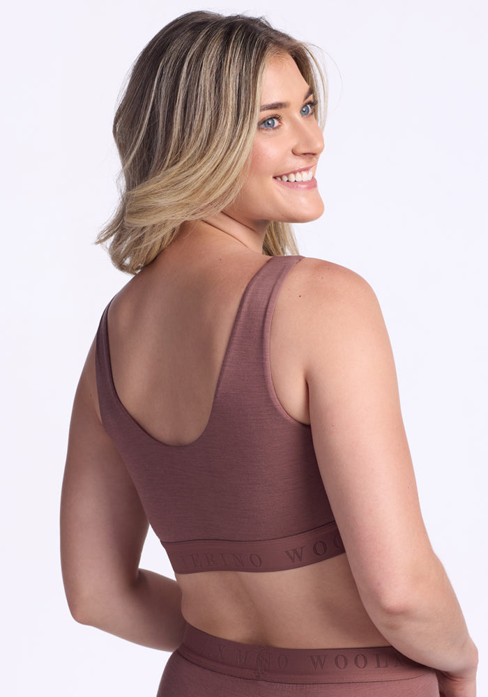 A woman with shoulder-length blonde hair smiles over her shoulder, wearing the Woolx Aubrey Lounge Bra in Nutmeg with a matching logo band bottom against a plain white background.