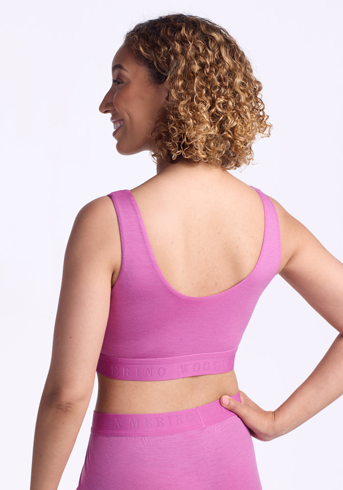 A woman with curly hair, seen from the back and smiling, models the Woolx Aubrey Lounge Bra in Wild Blossom with matching leggings against a plain light background, one hand resting on her hip.