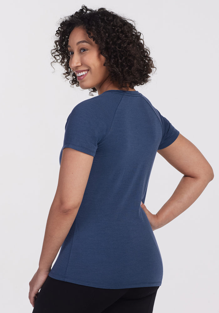 A woman with curly hair models the Woolx Mia V Neck - Starry Night, standing in black pants and smiling over her shoulder against a white background.