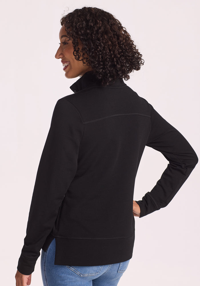 A woman with curly hair, wearing Woolx's Annie 3 Button Pullover in black, stands smiling with her back to the camera and right hand on her hip, looking over her shoulder.