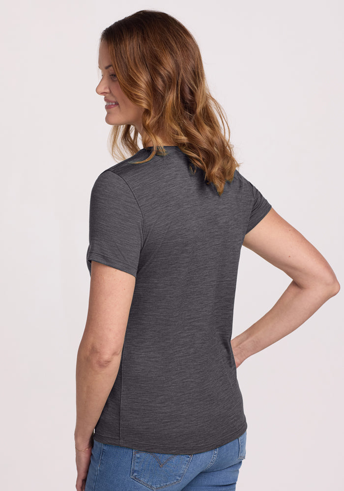 A woman with wavy brown hair, wearing the Woolx Liza Crew Neck Tee in Cinder Melange and blue jeans, stands with her back to the camera and one hand on her hip against a plain light background.