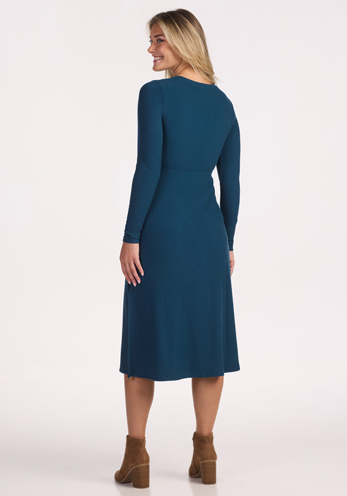 A woman with long blonde hair, wearing the Woolx Caroline Ribbed Twirl Dress in Majolica Blue and brown ankle boots, stands facing away and smiles over her shoulder against a plain light background.