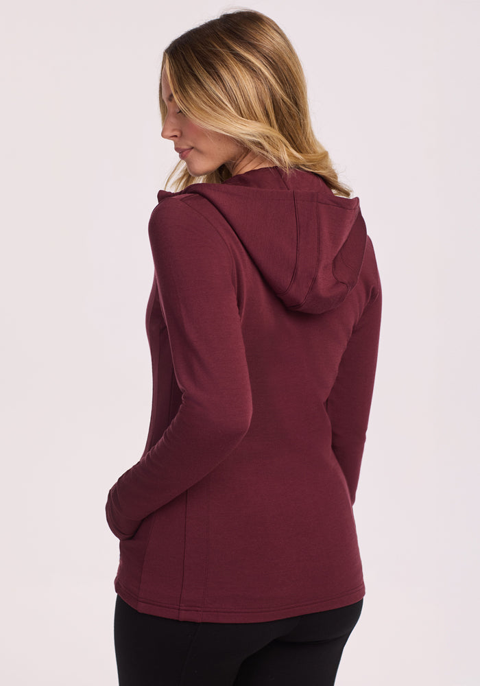 A woman with wavy blonde hair, facing away, wears the Woolx Zoey Zip Up Hoodie in Toasted Garnet with her hands in the pockets, standing against a plain light background.