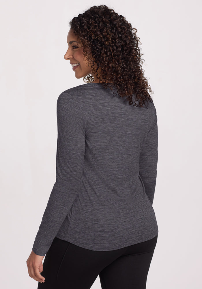 A woman with curly hair stands smiling over her shoulder against a plain light background, wearing the Woolx Millie Long Sleeve in Cinder Melange and black pants.