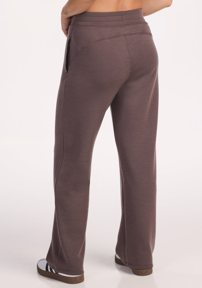A person wears Woolx Bree Lounge Pants in Plum Truffle, featuring a relaxed fit, straight legs, seam details, and side pocket zippers. The shot shows only the lower back and legs against a white background.