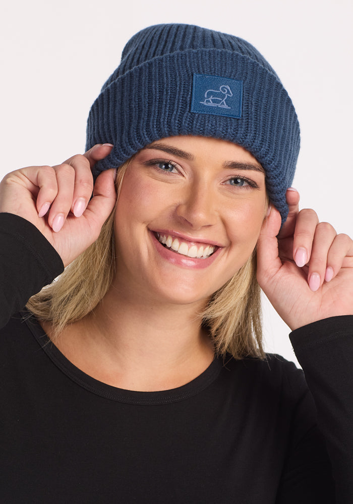 A smiling woman wears the Woolx Ria Chunky Beanie - Starry Night with a patch and a black long-sleeve shirt, holding the cozy winter accessory’s sides with both hands against a plain background.