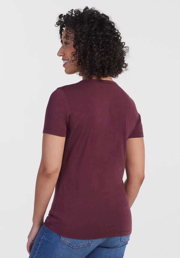 A person with short curly hair wears the Woolx Liza Crew Neck Tee in Grape Harvest and blue jeans, standing with their back to the camera against a plain light background.