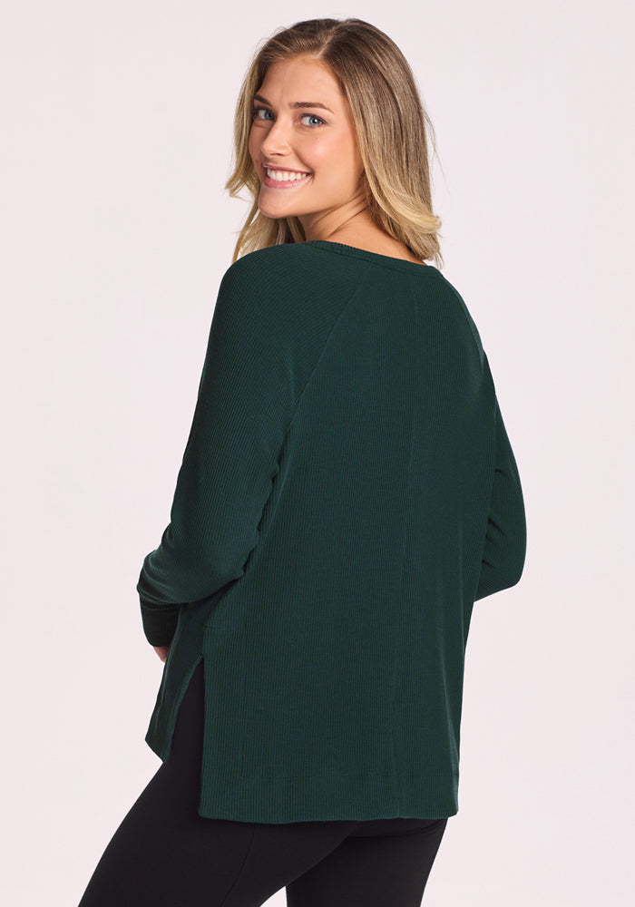 A woman with long, wavy blonde hair smiles over her shoulder, wearing the Woolx Ainsley Sweater in Deep Timber and black pants, standing against a plain light background.