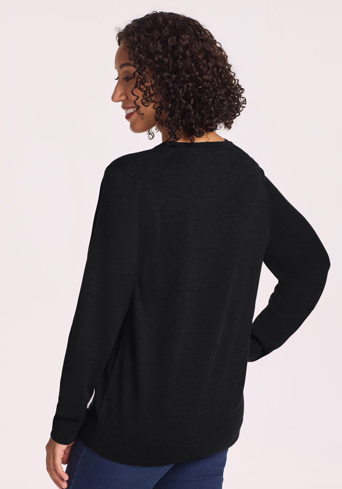 A woman with curly hair wears the Woolx Francesca Sweater in black and blue jeans, standing with her back to the camera and smiling over her shoulder against a light background. This travel-friendly sweater offers effortless style.