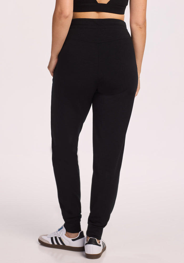 A person wearing Woolx Willa Joggers in black is shown from the waist down, paired with a black crop top and white sneakers with black stripes, facing away from the camera against a plain light background.