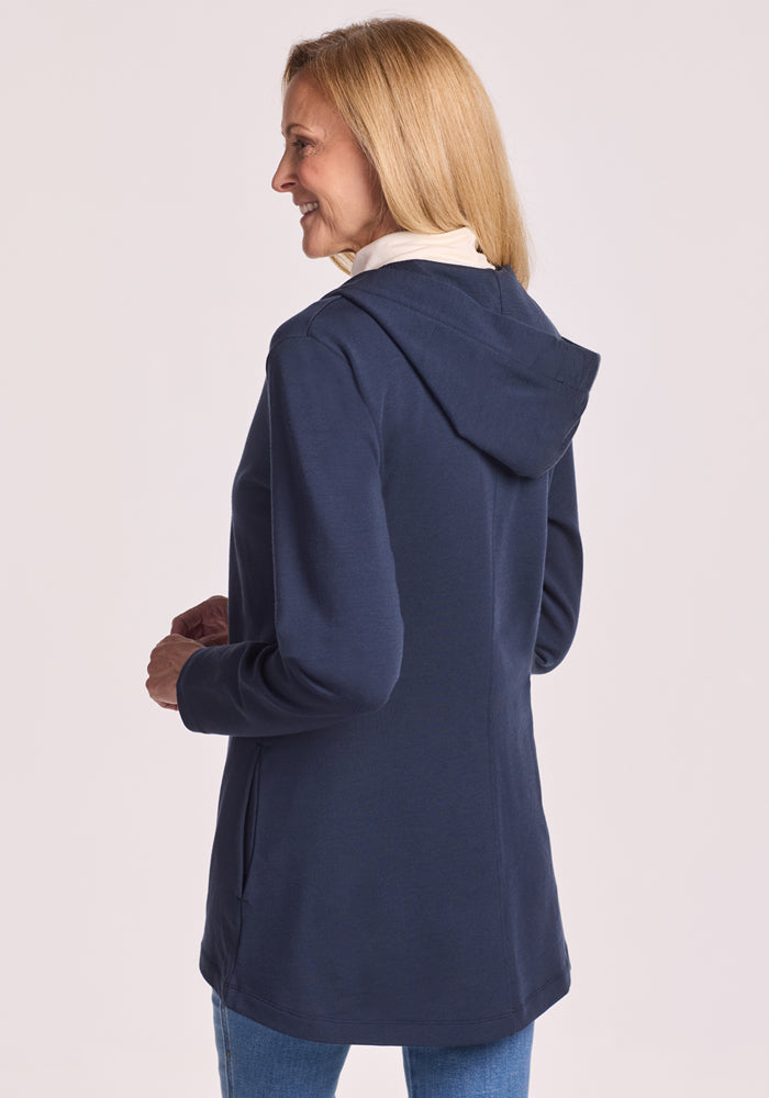 A woman with long blonde hair, wearing the Woolx Edna Hooded Cardigan in Deep Navy over a cream top and blue jeans, stands smiling with her back to the camera—ready for any cold-weather outing.