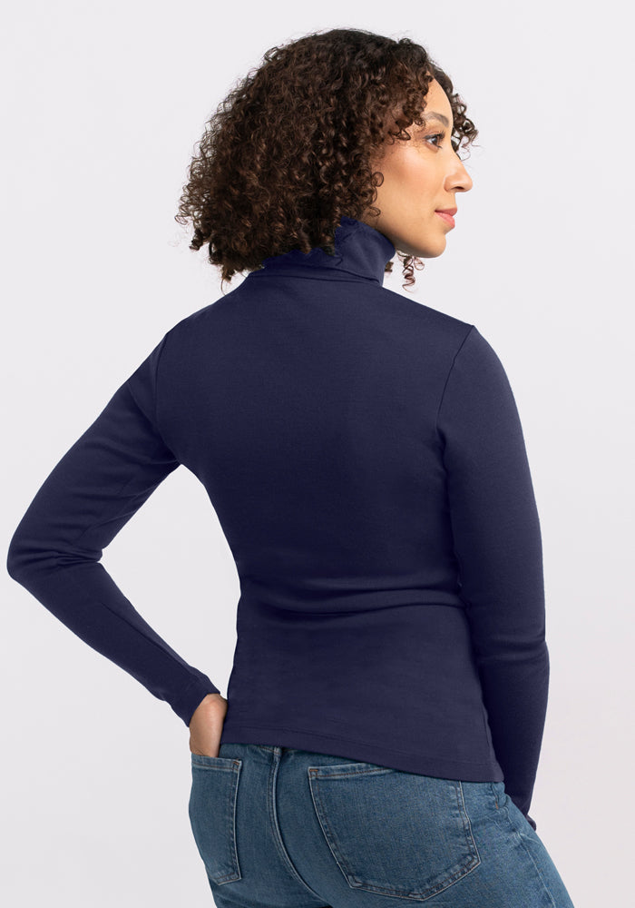 A woman with curly hair models the Woolx Peyton Turtleneck in Midnight Sapphire and blue jeans, standing with her back to the camera and glancing over her shoulder against a plain light background.