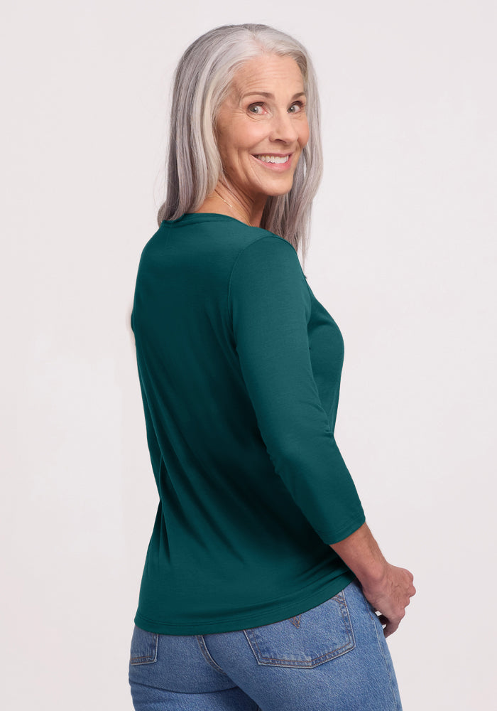 A smiling older woman with long gray hair stands against a plain light background, wearing blue jeans and the Woolx Jenny 3/4 Sleeve Crew Neck in Riverbank Green, looking over her shoulder.