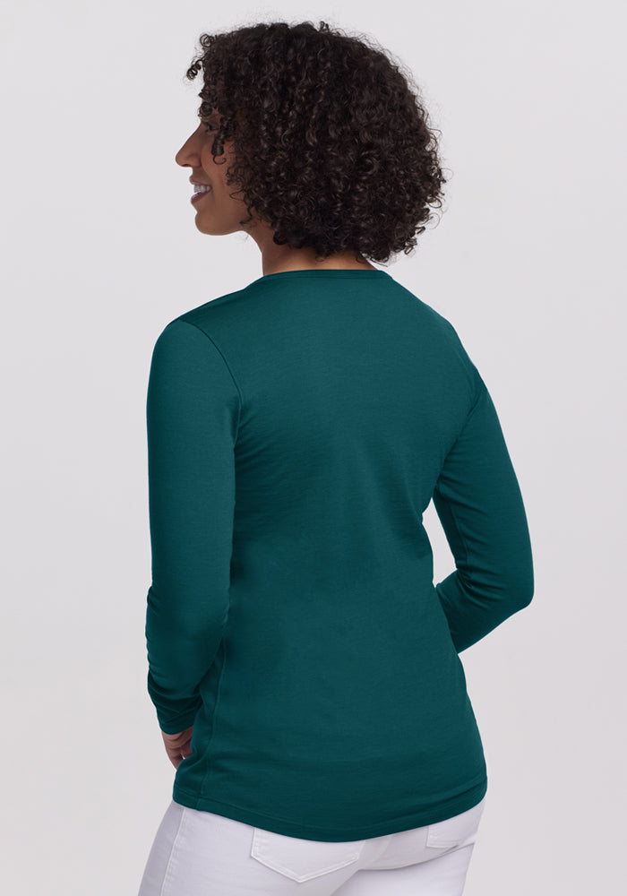 A woman with curly hair, wearing the Woolx Remi Long Sleeve T-Shirt in Riverbank Green and white pants, stands facing away from the camera and smiles over her shoulder against a plain light background.